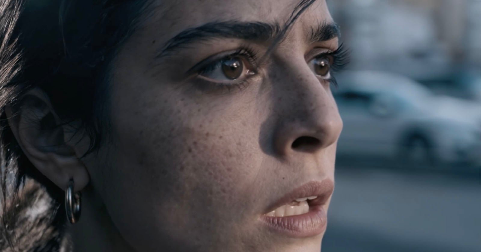A close-up of a woman with dark hair and brown eyes, looking ahead with a worried or anxious expression. Her mouth is slightly open, and the background appears blurred, showing an outdoor scene.