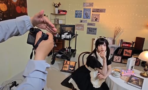 A person in a maid costume poses at a vintage-themed table while another person takes a photo. The background has retro posters, books, and soft lighting, creating a cozy, nostalgic atmosphere.