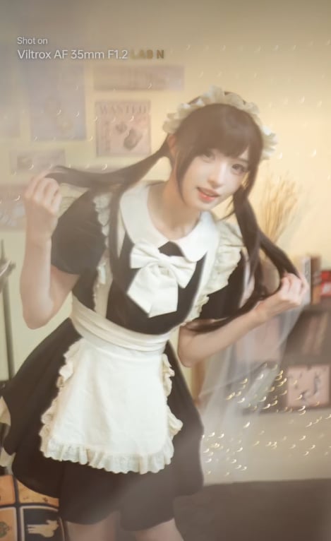 A person wearing a black and white maid outfit with a frilly apron and headband poses indoors, playfully holding their pigtails. The background is softly lit with decorative lights and blurred shelves.
