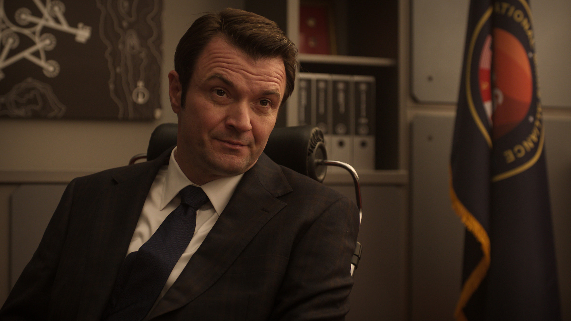 a man in a suit sits behind a desk in an office