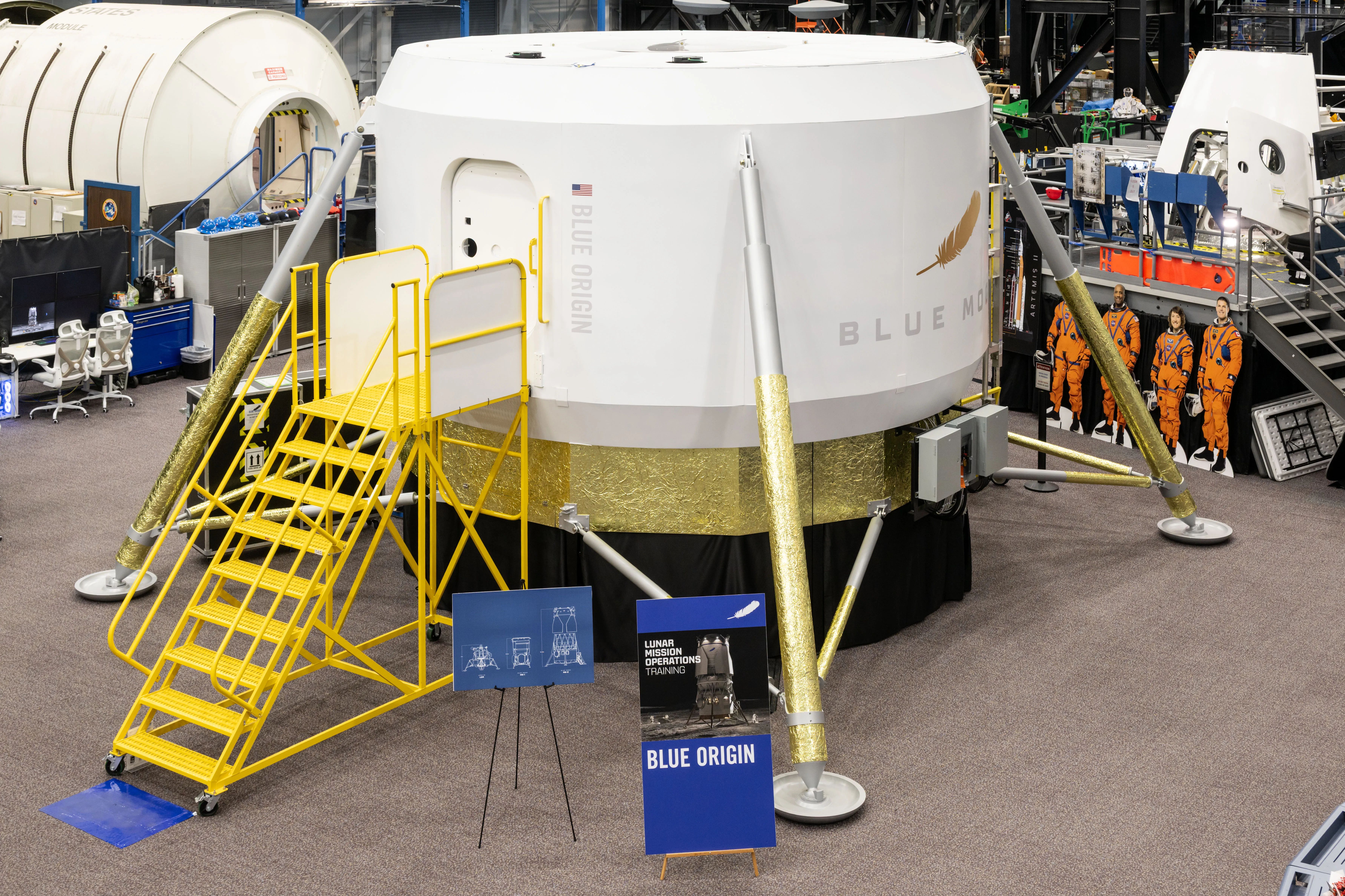 a cylindrical white capsule stands on four golden-foil-wrapped legs with yellow scaffolding steps against a rectangular hatch with rounded corners inside a big spacecraft mockup training facility.
