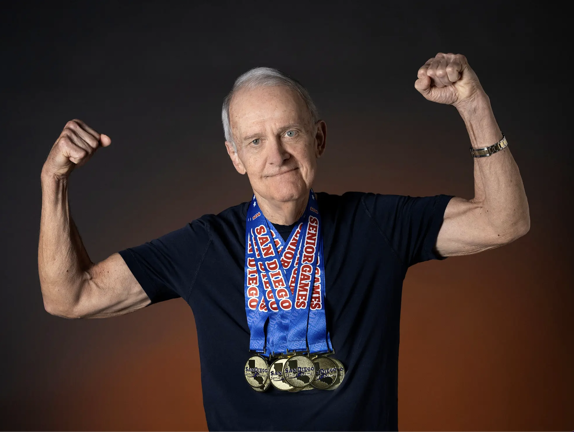 Senior man in biceps flexing pose, wearing multiple gold medals with "San Diego Senior Games" written on the neck ribbons