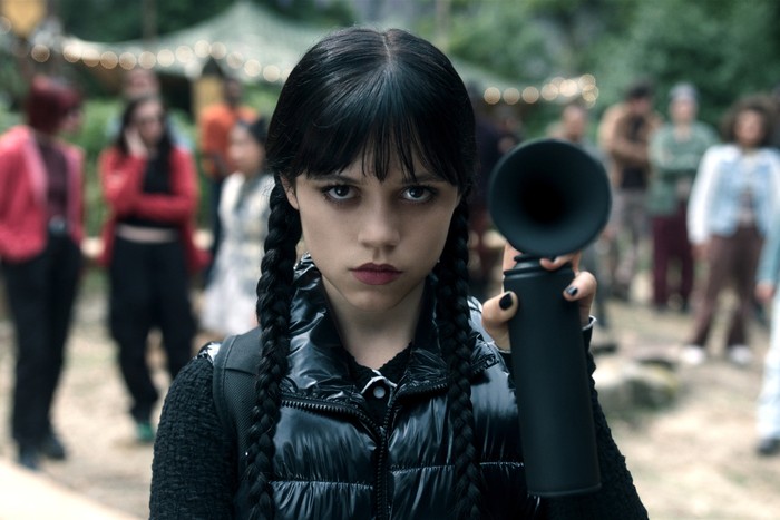 Wednesday-Netflix-S2-JennaOrtega Jenna Ortega as Wednesday Addams in Wednesday holding up a black horn as she stands outside amongst a crowd of people with a serious expression on her face.