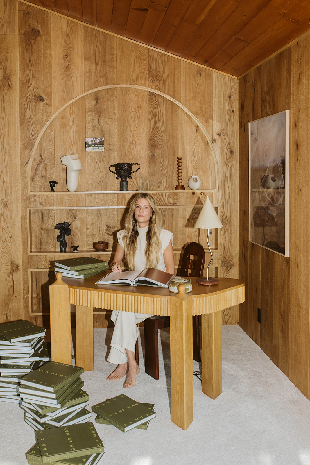 Portrait of a young blonde woman with long hair, dressed in casual-chic clothes in a studio while sitting on a wood and leather chair, or in a wood-clad office surrounded by books.