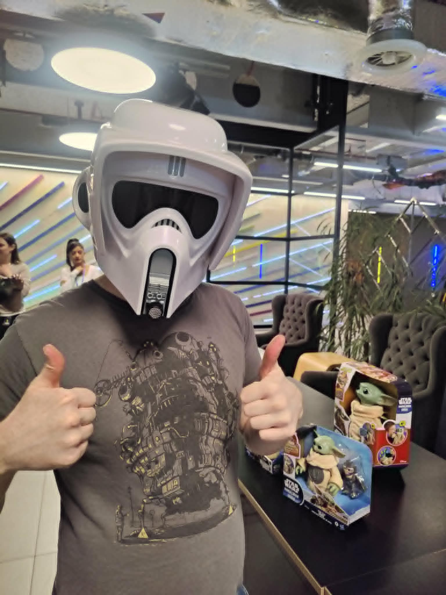 A man stands in a well-lit office giving a thumbs up to a camera while wearing a stormtrooper helmet.