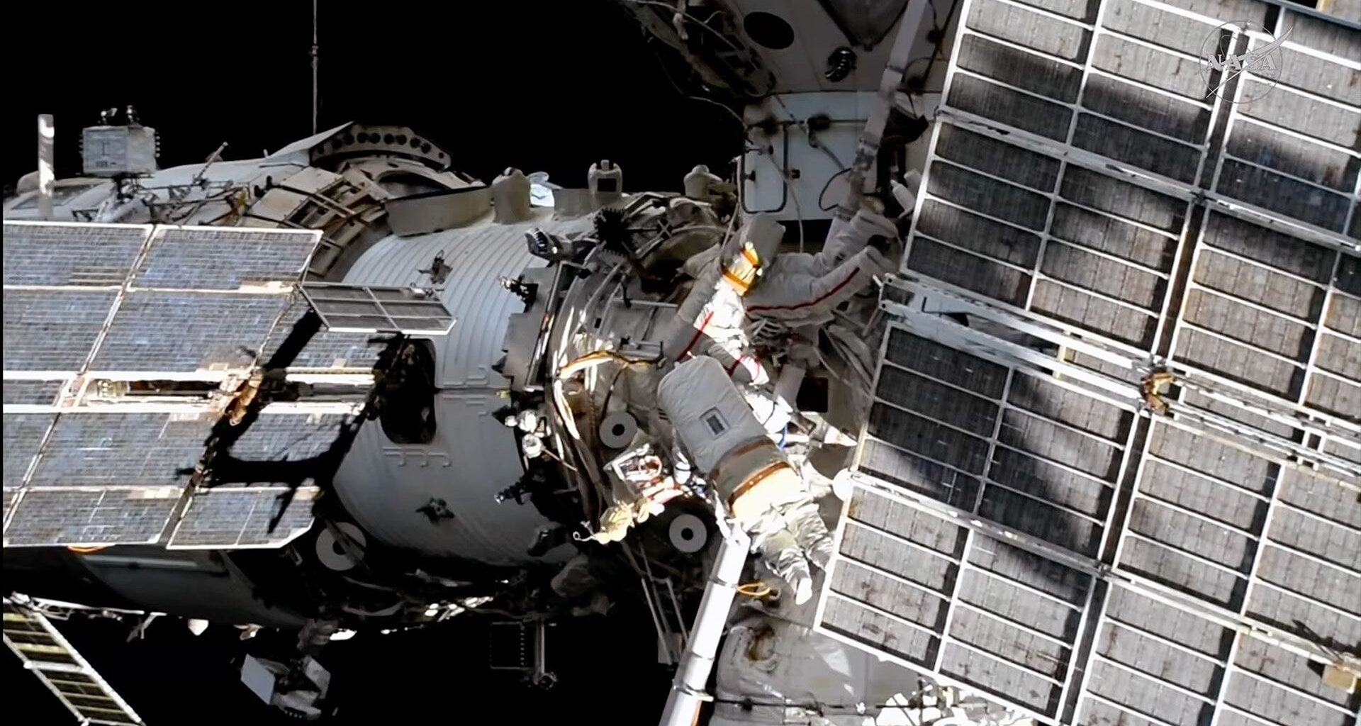 Two spacesuited cosmonauts are seen working outside a large space station set against the blackness of space.