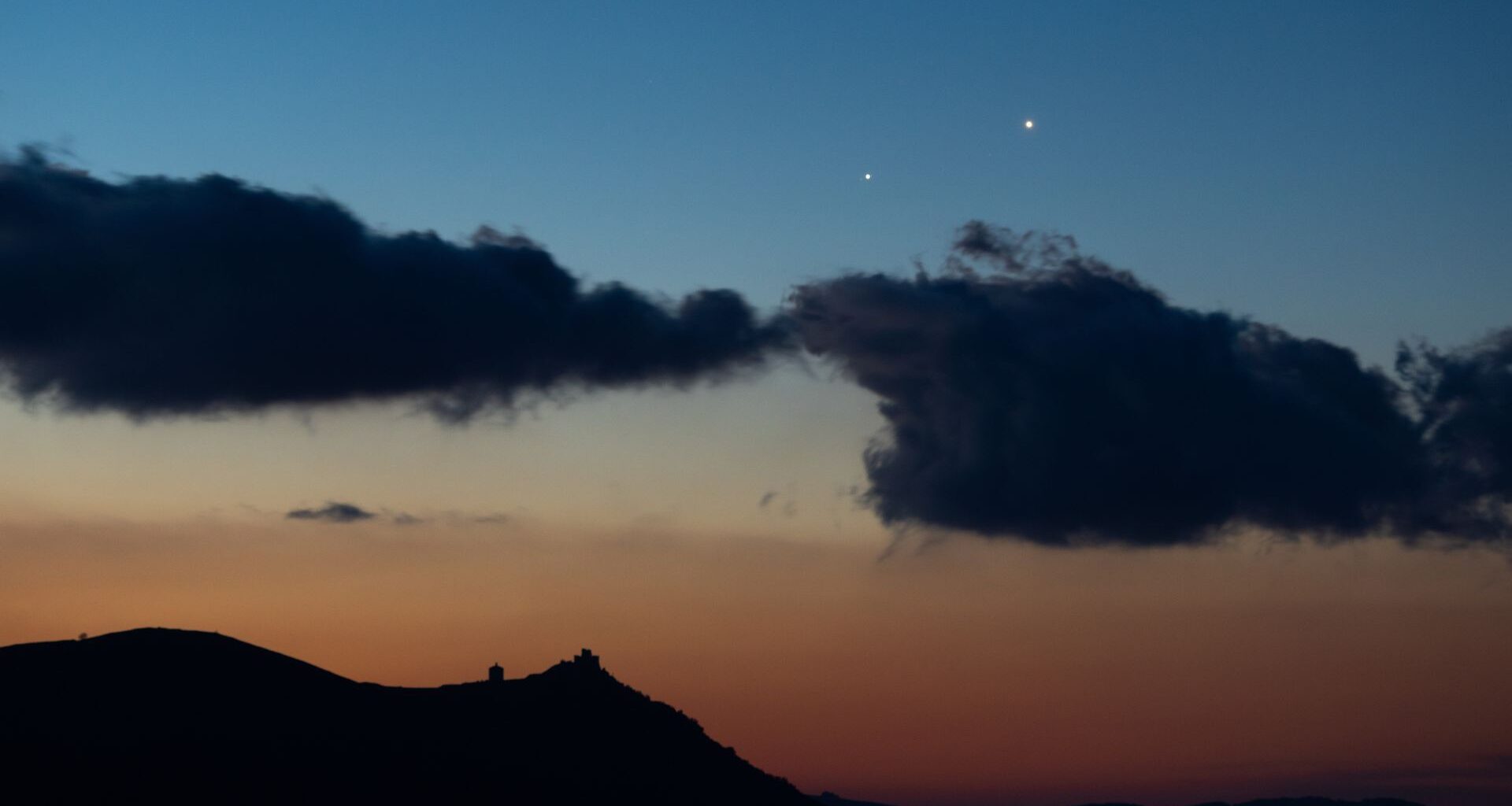 Two star-like planets shine close to one another in the evening sky above the silhouettes of clouds.