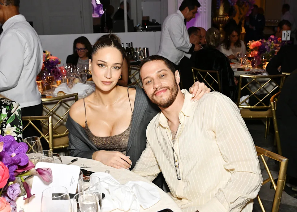 A man and a woman seated together at an elegant event, surrounded by tables and guests. The woman wears a low-cut top, and the man wears a light shirt
