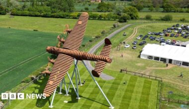 An aerial view of a large, brown‑coloured metal sculpture shaped like a plane mounted on angled support legs in the middle of a neatly mown field. The surrounding area includes open green fields, a curving path and a rural landscape stretching into the distance under a cloudy sky. To the right, a grassy car park filled with vehicles and a long white marquee suggests an outdoor event, with several people walking around the site.