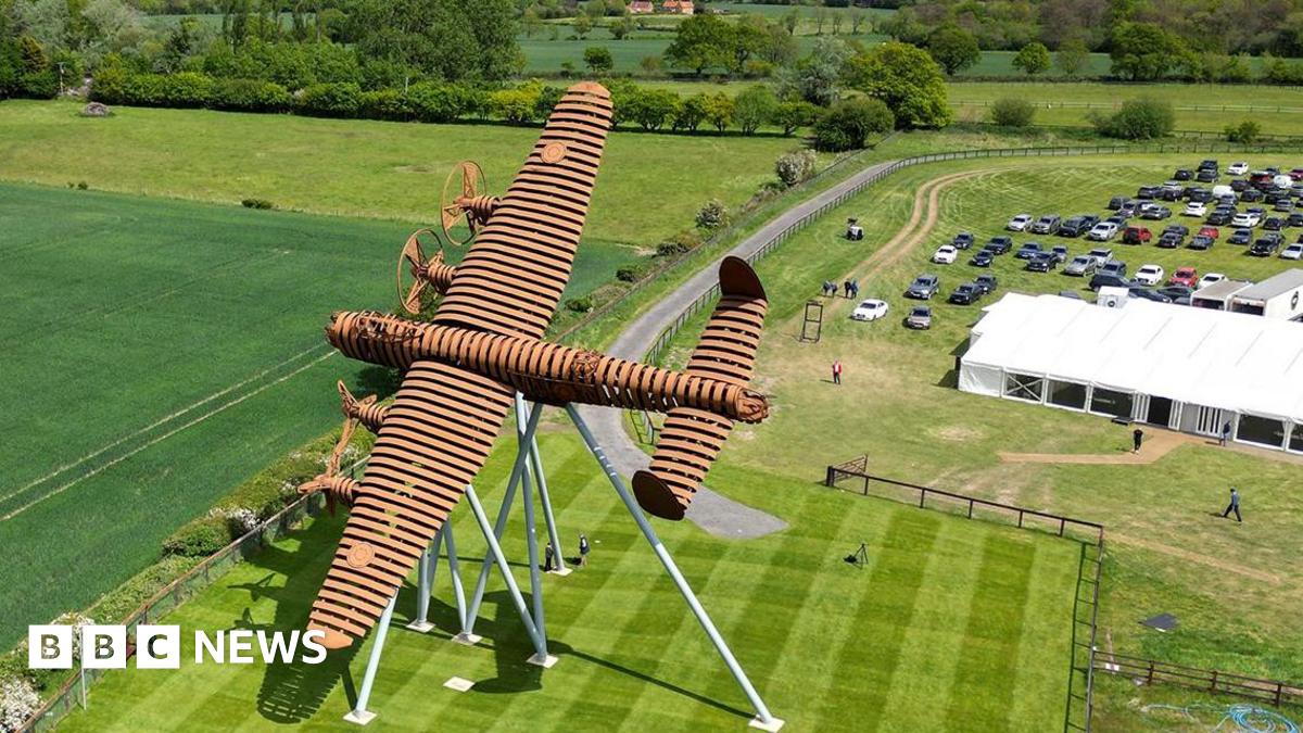 An aerial view of a large, brown‑coloured metal sculpture shaped like a plane mounted on angled support legs in the middle of a neatly mown field. The surrounding area includes open green fields, a curving path and a rural landscape stretching into the distance under a cloudy sky. To the right, a grassy car park filled with vehicles and a long white marquee suggests an outdoor event, with several people walking around the site.