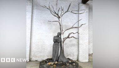 A statue including a shiny metal tree without leaves and a person leaning up against it on a scenic base with a white brick wall behind it