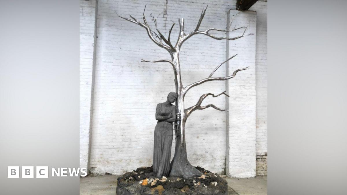 A statue including a shiny metal tree without leaves and a person leaning up against it on a scenic base with a white brick wall behind it