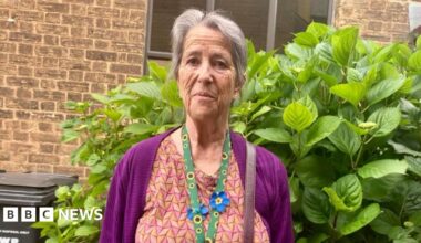 A woman in her 60s with long grey hair is wearing a purple cardigan and orange patterned dress. She is standing in her garden.