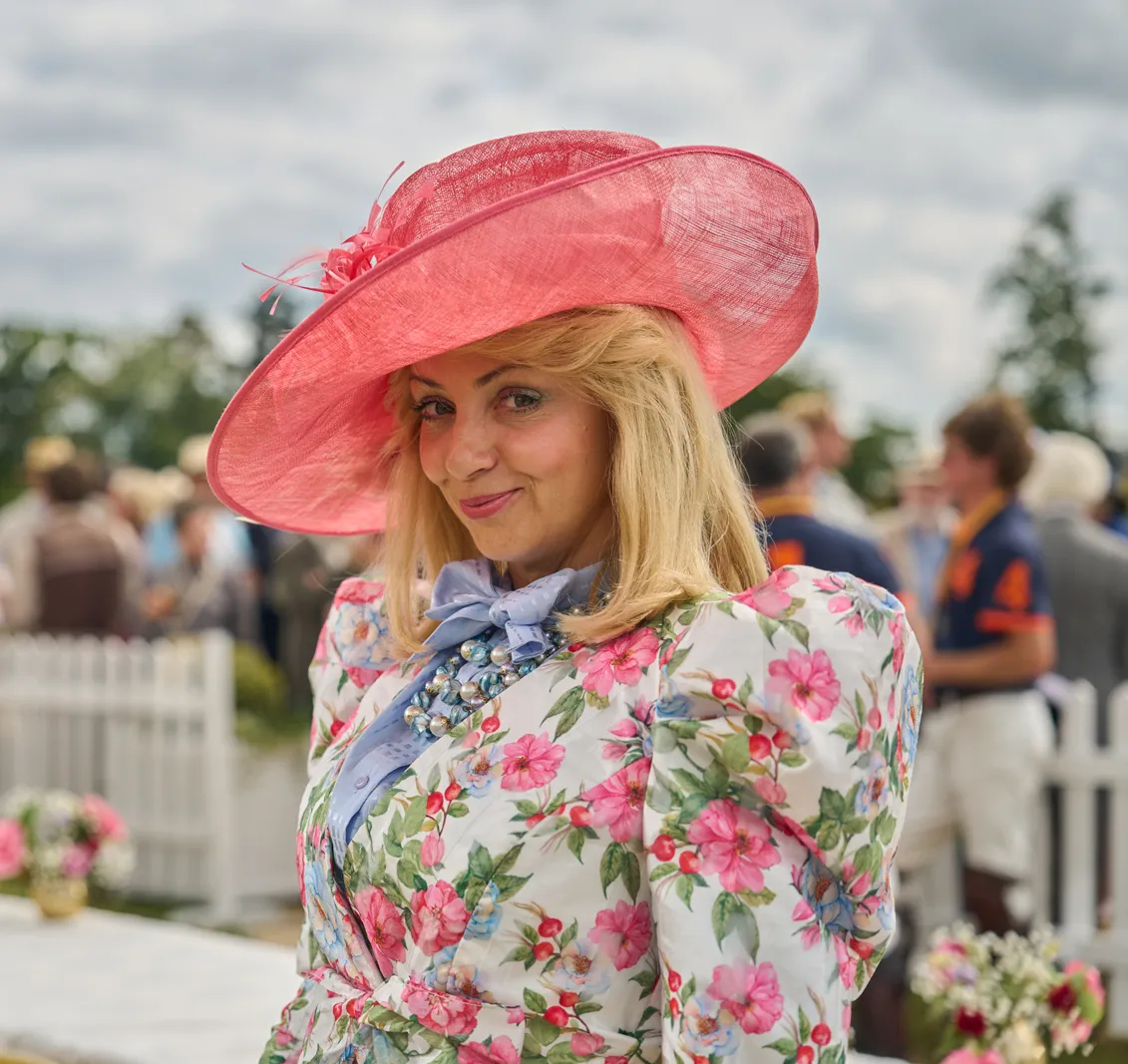 A woman in a wide-brimmed pink hat, floral dress, and light blue bow tie smiles, with a polo match in the background.