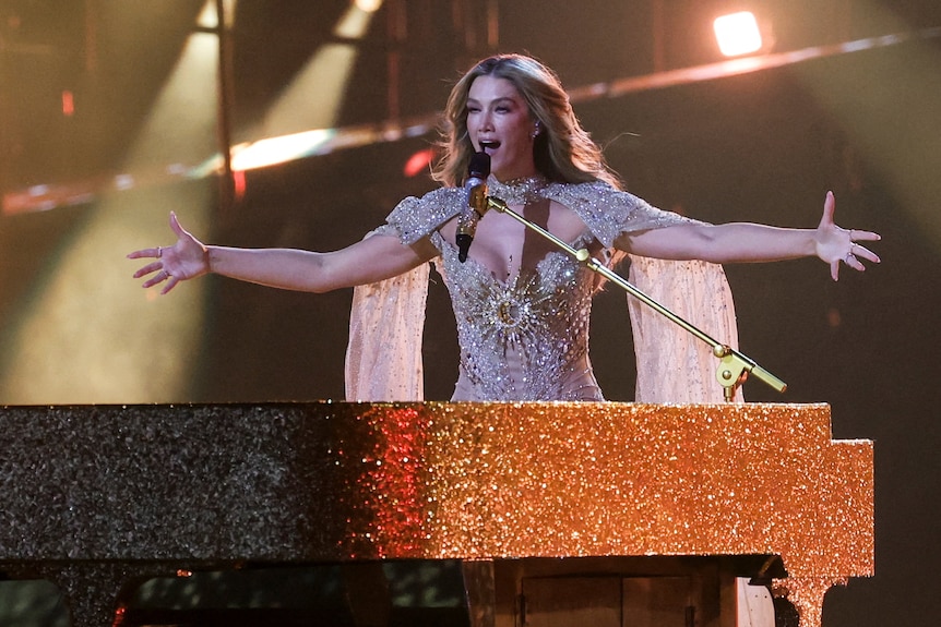 Delta Goodrem with her arms outstretched standing behind a golden piano.