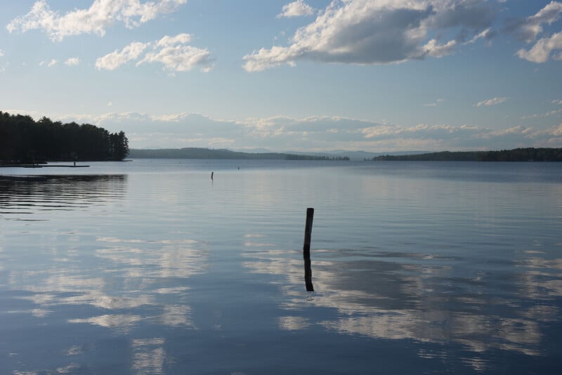 A calm lake under a partly cloudy sky, with trees lining the distant shore and a wooden post sticking out of the water, reflecting in the gentle ripples.