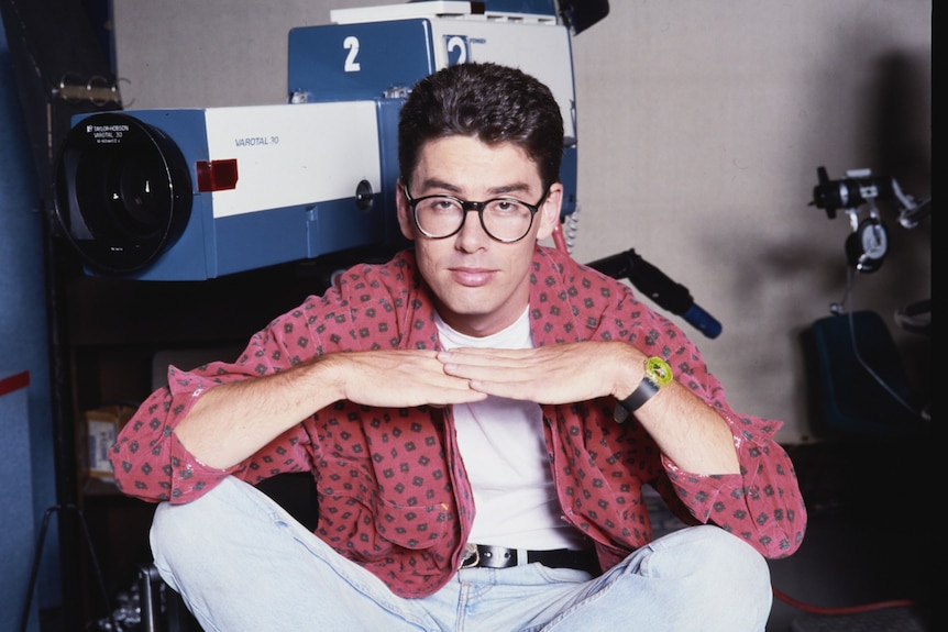 Man with brown hair and thick-rimmed glasses sits cross-legged on the floor, his hands under his chin