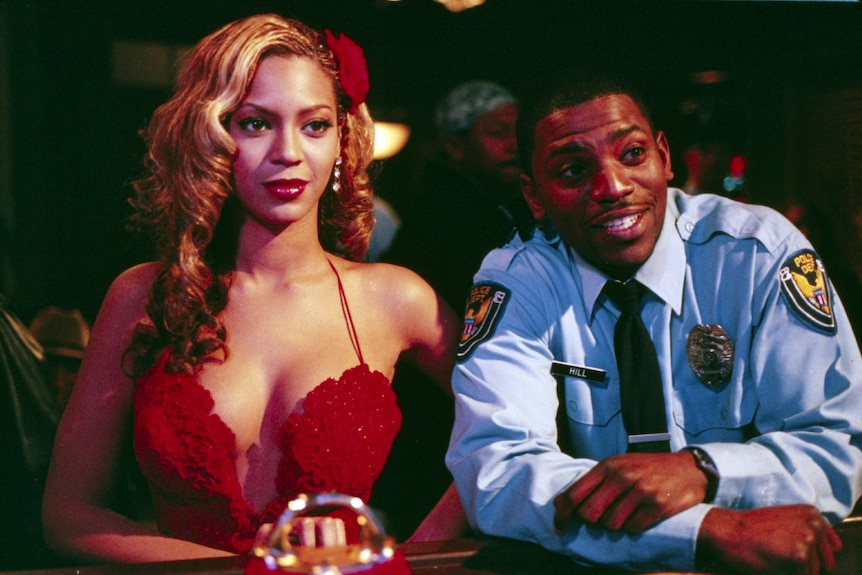 Beyoncé as Carmen in a red dress and red rose in hair, standing at the bar next to Sgt Hill in full uniform