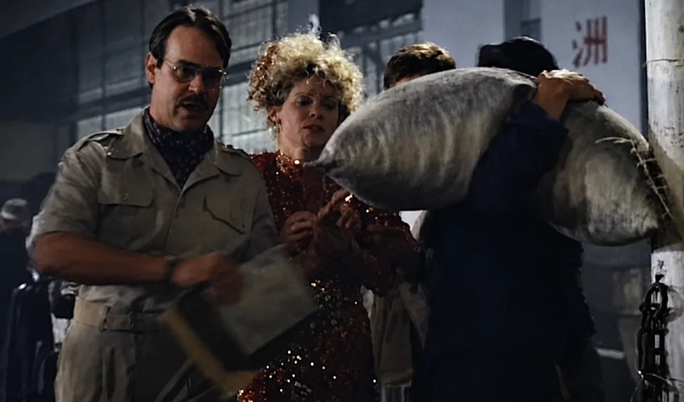 A group of people stand together, one holding a large sack. A person in a sparkly dress looks on. They appear to be on a movie set or film scene