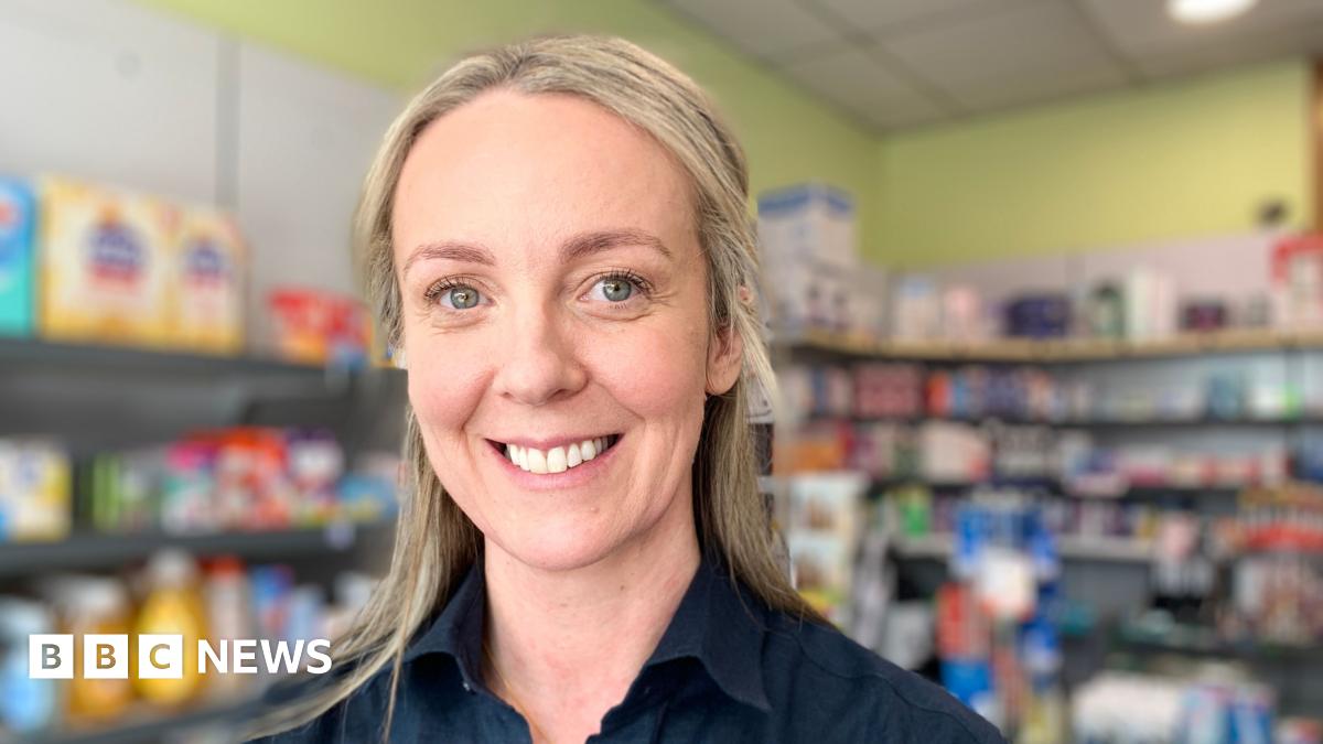 Two Female Pharmacists Working at a Pharmacy. They are wearing white coats and there are shelves filled with medication in the background.