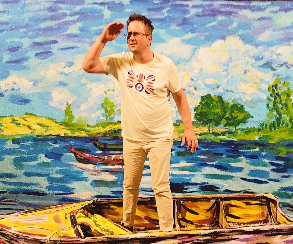 A man in light-colored clothing and a graphic tee stands inside a painted boat, posing with his hand to his forehead against a vibrant, impressionistic lake backdrop.