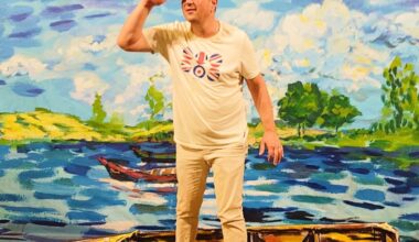 A man in light-colored clothing and a graphic tee stands inside a painted boat, posing with his hand to his forehead against a vibrant, impressionistic lake backdrop.