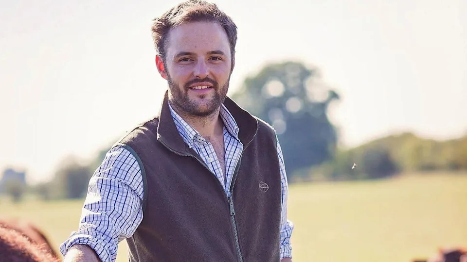 James Florey is wearing a shirt under a brown gilet and is touching a cow. He is standing outside in a field.