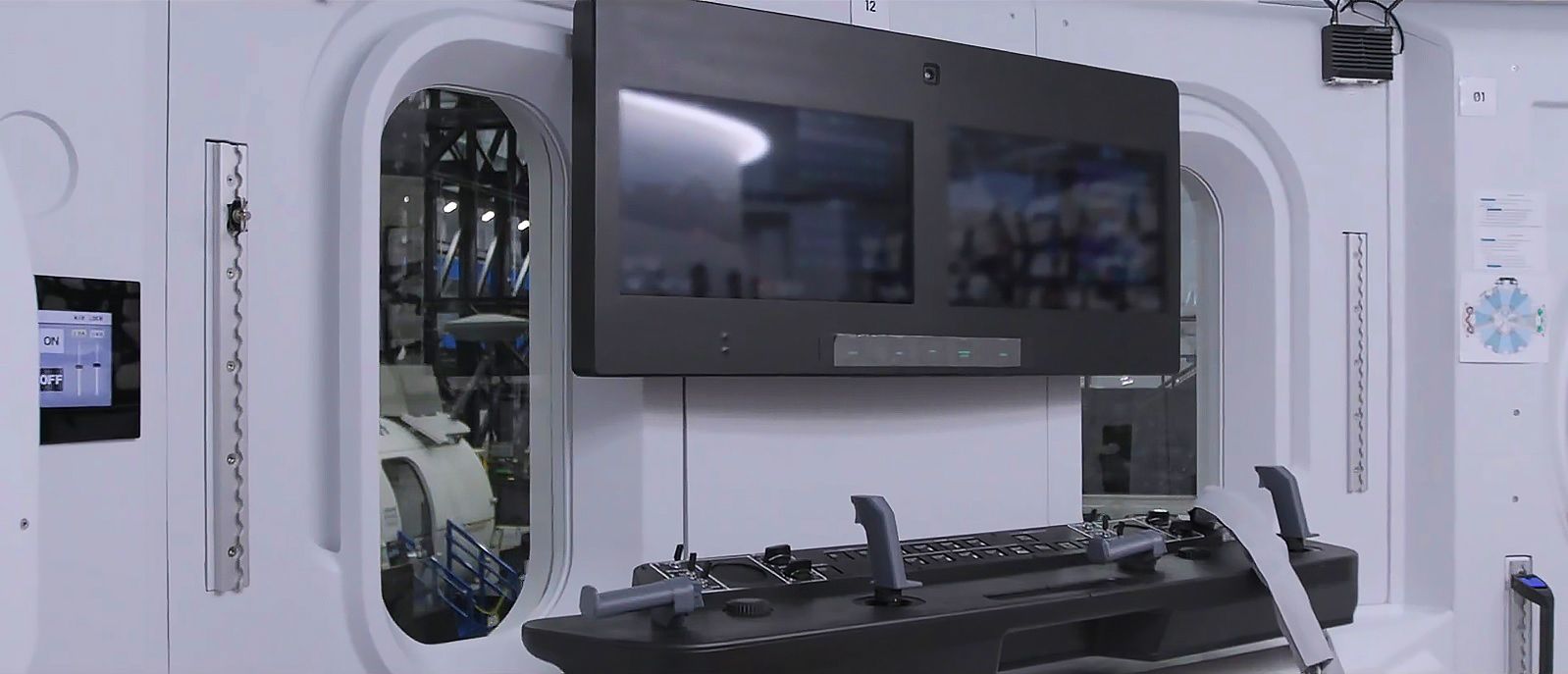 a joystick instrument panel sits under two black screens, with two tall oval windows on either side, inside a white room.