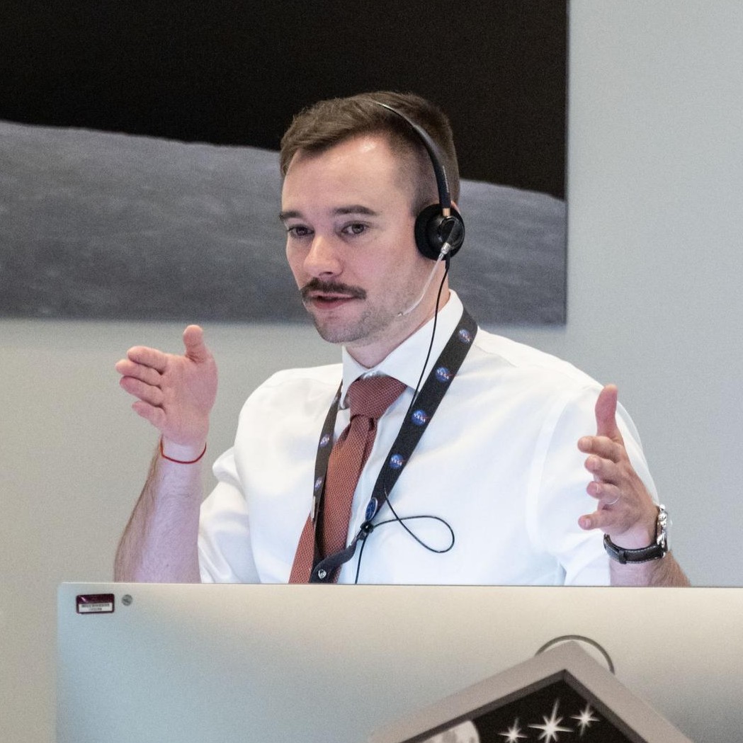 a man with a mustache stands, looking focused, with his hands spread apart as if showing you the size of something.