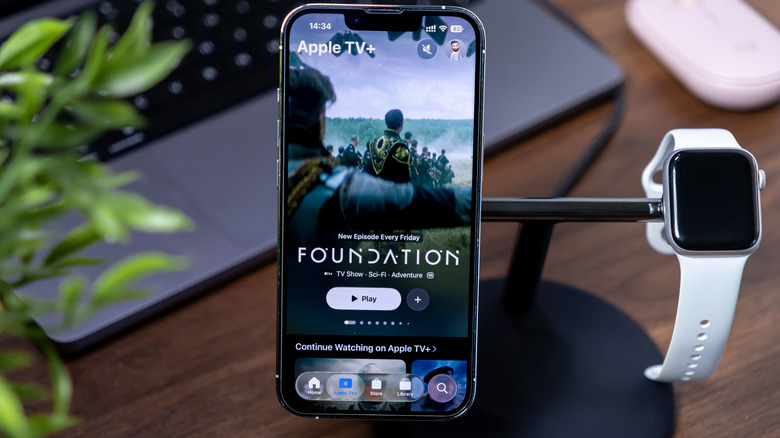pple TV app showing Foundation series on an iPhone with iOS 26 and Liquid Glass, with Apple Watch charging on a modern desk setup