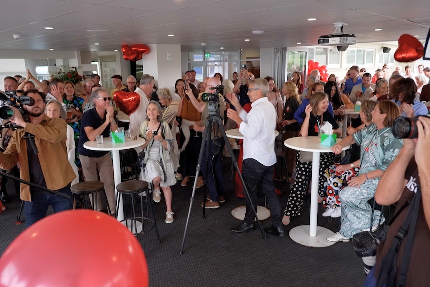 A crowd of people, many clapping, with red heart-shaped balloons visible.