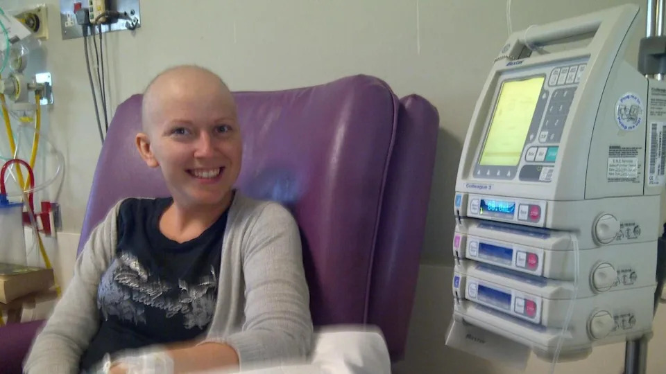 A woman sat in a chair undergoing cancer treatment. She is smiling at the camera. There is hospital machinery to her left, our right