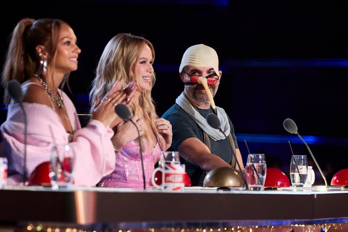 Alesha Dixon, Amanda Holden and Simon Cowell on Britain's Got Talent Simon Cowell wearing a bizarre bald cap and long thin nose, with red-tinted glasses on, sitting on the BGT panel alongside Alesha Dixon and Amanda Holden, who are smiling at what they're watching.