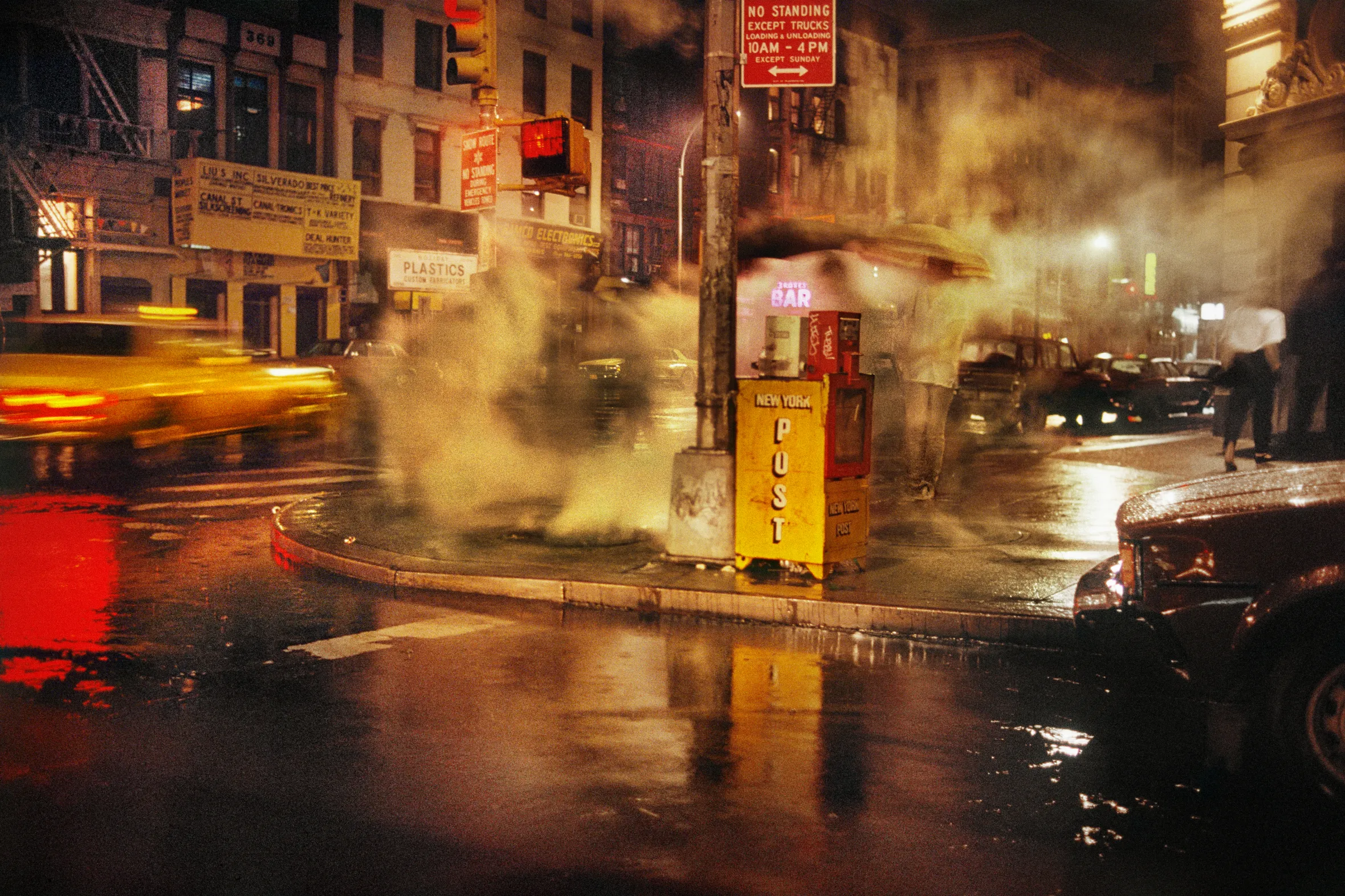 A rainy, smoky street corner in Manhattan at night with a bright yellow taxi driving past.