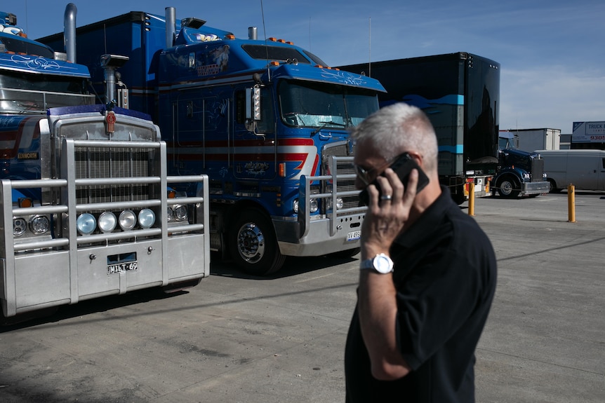 A man in a black polo shirt speaks on his phone in front of two massive blue and red trucks.