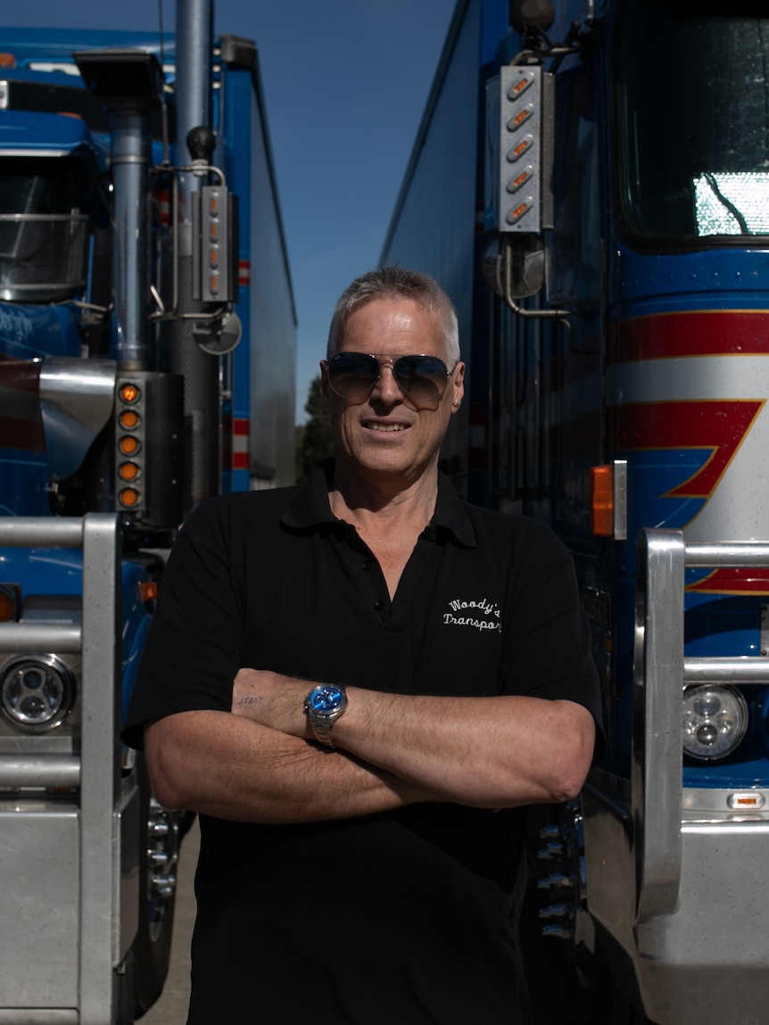 A man in a black polo and sunglasses stands with his arms crossed in front of two massive blue and red trucks.