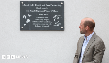 Prince William, wearing a grey suit and a light blue shirt. He is standing in front of a grey wall. He is looking to his right at the wall. There is a wall plaque which reads "Isles of Scilly Health and Care Partnership officially opened by His Royal Highness Prince William. 22 May 2026. A celebration of the Isles of Scilly Hospital and Islands View Care Home, delivered in partnership with the Duchy of Cornwall."