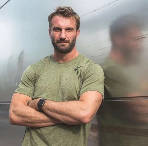 Luke Worthington, an adult man with a beard, wearing a green t-shirt, standing with arms crossed.