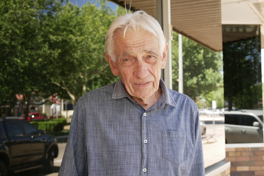 An older man with white hair and a blue shirt stands in front of a window frowning