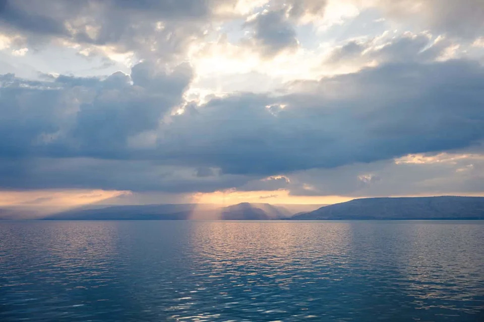 A serene view of the Sea of Galilee at sunset with sun rays breaking through dramatic clouds.