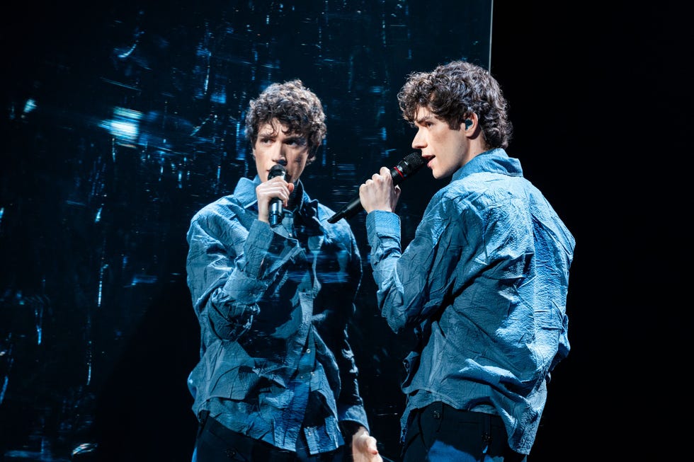 a performer with curly hair stands in front of a mirror while holding a microphone