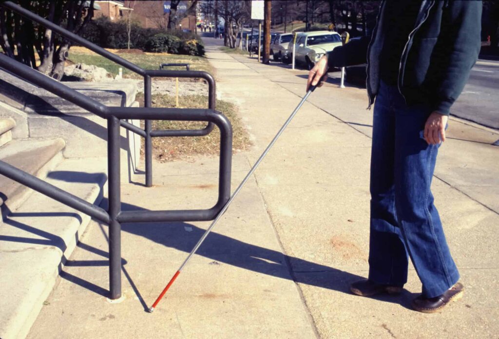Blind person with a white cane navigating the sidewalk near stairs.