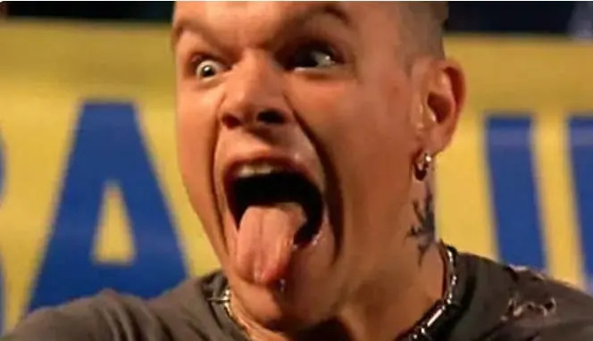A person with a surprised expression sticks their tongue out, wearing earrings and a neck tattoo, set against a blurred background