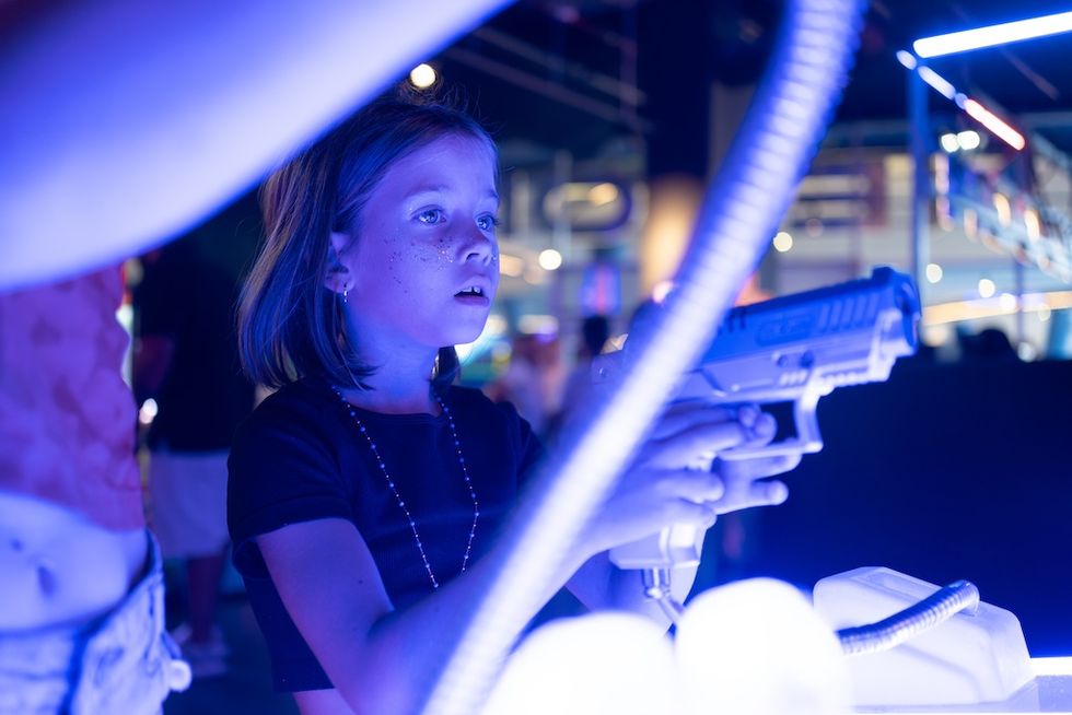 Child playing an interactive arcade game with a toy gun, illuminated by blue light.