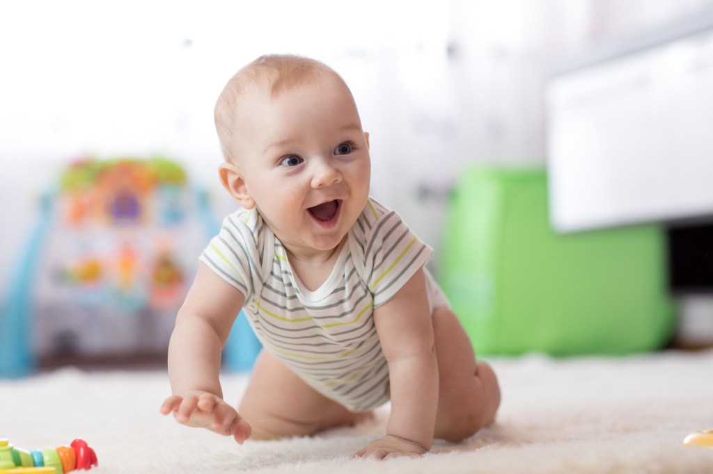 A happy baby boy in a striped onesie crawls on a white rug indoors.