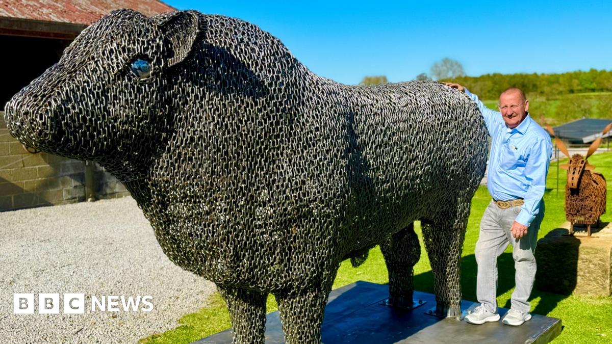 Steven Nesbitt  standing at his farm with his arm around his big metal bull.  He is wearing a light blue shirt and grey jeans, and has a shaved head. It is a bright sunny day.
