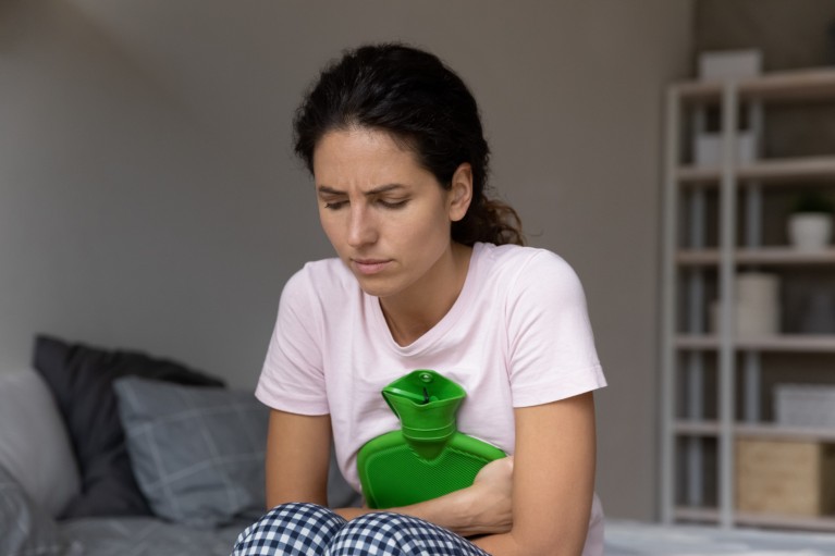 A woman in pyjamas sat on the edge of a bed, holding a hot water bottle to her stomach.