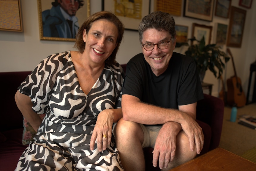 Man and a woman sit side-by-side on a large red couch. Walls covered in photo frames