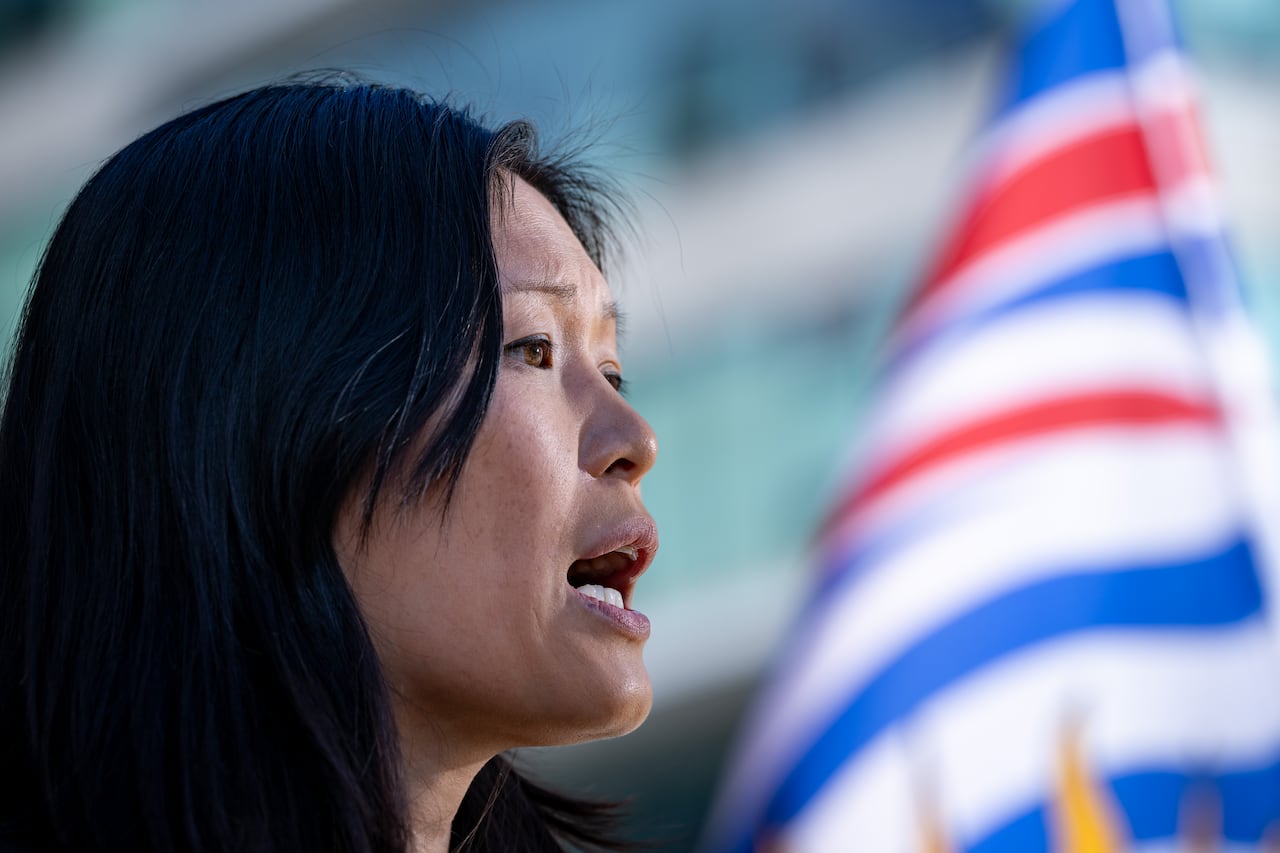 An East Asian woman speaks in front of a B.C. flag.