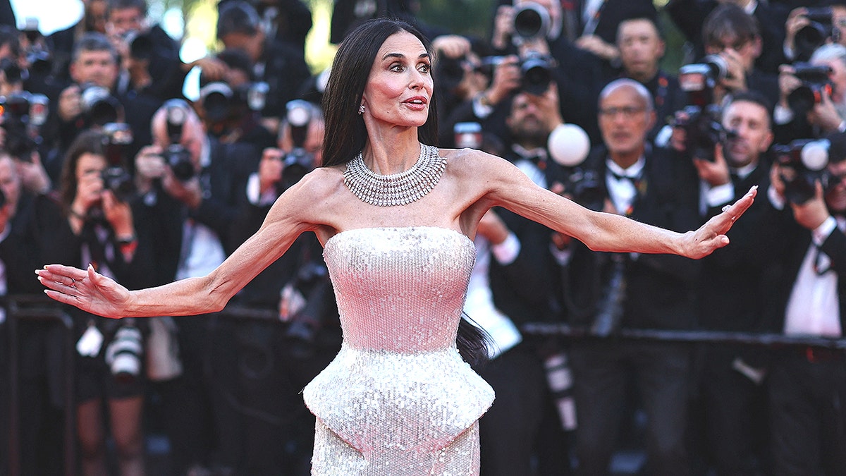 Demi Moore in a strapless shimmering gown poses with arms outstretched on a red carpet while photographers crowd behind her taking pictures.